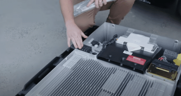 Person handling a large, gray electronic device with internal components exposed, including circuit boards and cables, placed on the ground.